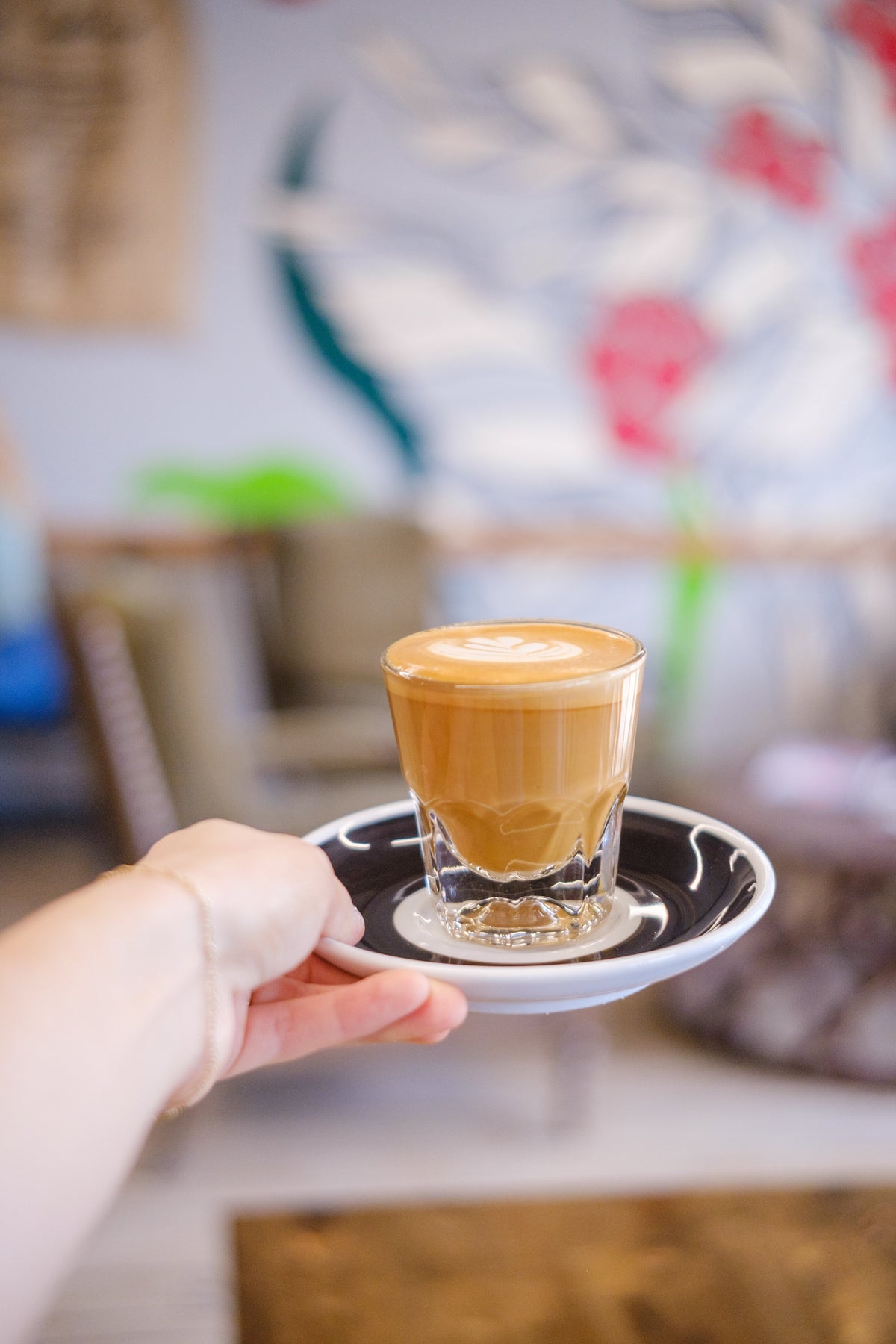 female hand holding a cortado with a painted coffee plant blurred in the background